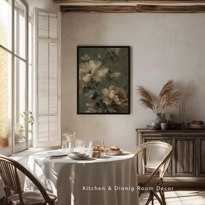 Op de afbeelding: Een ingelijste bloemenprint met cr&egrave;mekleurige en bruine bloemen tegen een gedempte groene achtergrond hangt boven een eettafel. De tafel is gedekt met witte borden en glazen. De tekst "Kitchen & Dining Room Decor" staat onderaan.