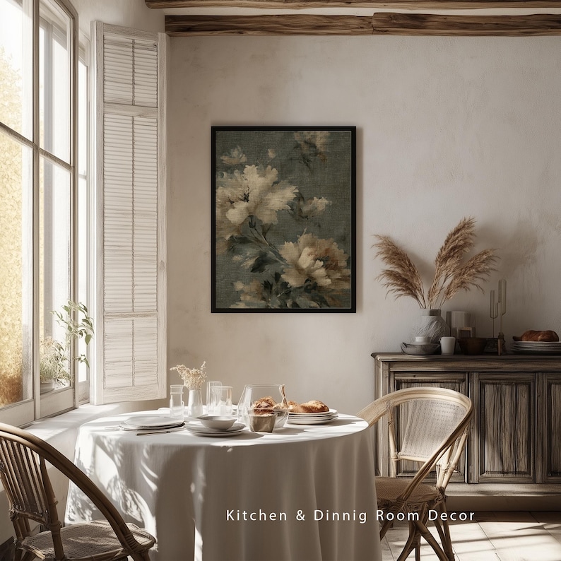 Op de afbeelding: Een ingelijste bloemenprint met cr&egrave;mekleurige en bruine bloemen tegen een gedempte groene achtergrond hangt boven een eettafel. De tafel is gedekt met witte borden en glazen. De tekst "Kitchen & Dining Room Decor" staat onderaan.