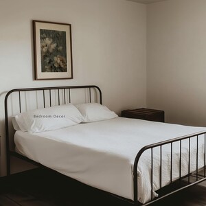 Op de afbeelding: Een slaapkamer met een zwart metalen bedframe, witte beddengoed en kussens. De kussens hebben de tekst "Bedroom Decor". Een ingelijste bloemenprint hangt boven het bed. Een donkere houten kist staat naast het bed.