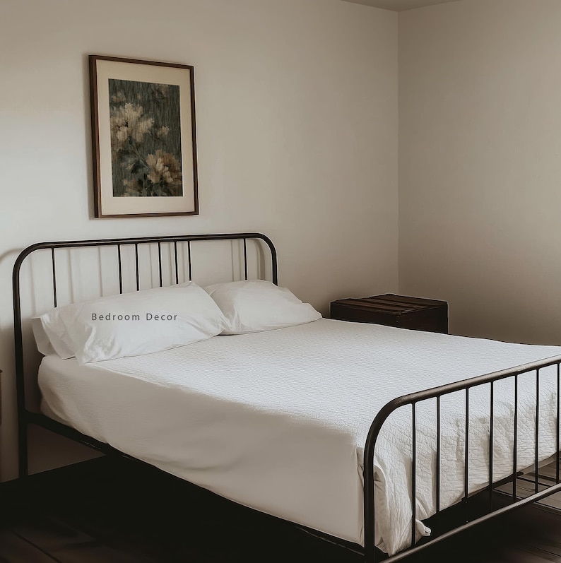 Op de afbeelding: Een slaapkamer met een zwart metalen bedframe, witte beddengoed en kussens. De kussens hebben de tekst "Bedroom Decor". Een ingelijste bloemenprint hangt boven het bed. Een donkere houten kist staat naast het bed.