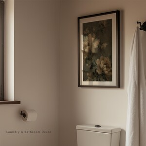 Op de afbeelding: Een ingelijste bloemenprint met een donkerbruine lijst hangt aan een lichtbeige muur in een badkamer. Een witte handdoek hangt aan een zwarte haak. Een rol toiletpapier is aan de muur bevestigd. De tekst "Laundry & Bathroom Decor" staat onderaan.