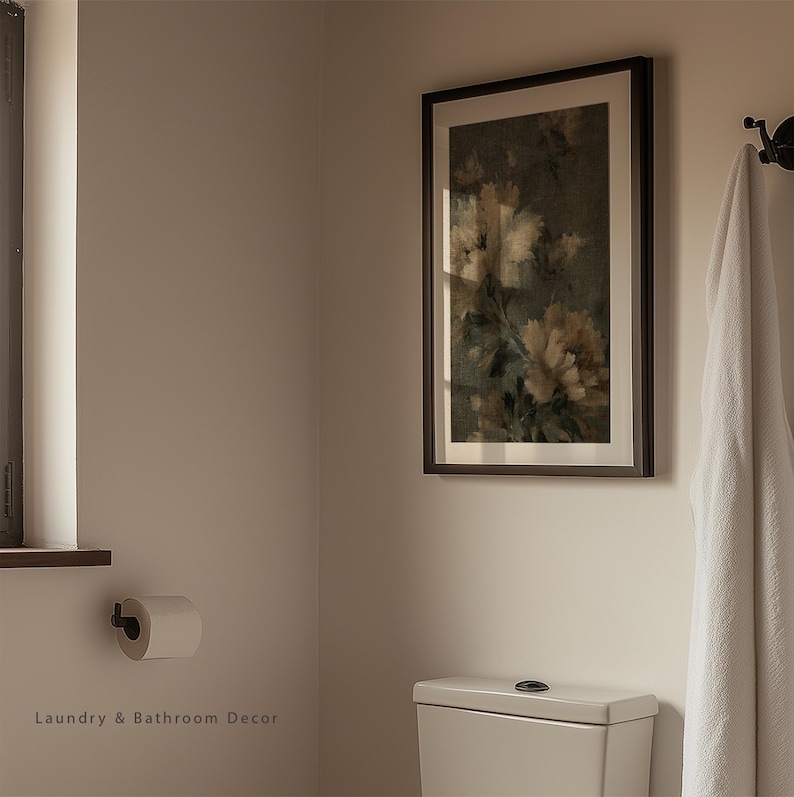Op de afbeelding: Een ingelijste bloemenprint met een donkerbruine lijst hangt aan een lichtbeige muur in een badkamer. Een witte handdoek hangt aan een zwarte haak. Een rol toiletpapier is aan de muur bevestigd. De tekst "Laundry & Bathroom Decor" staat onderaan.