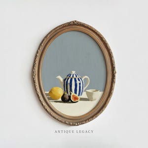Può includere: Un dipinto a olio in cornice ovale raffigurante una teiera a righe blu e bianche, un limone giallo, un fico scuro e una piccola tazza bianca su una superficie chiara con sfondo grigio. Il testo "ANTIQUE LEGACY" è stampato sotto la cornice.