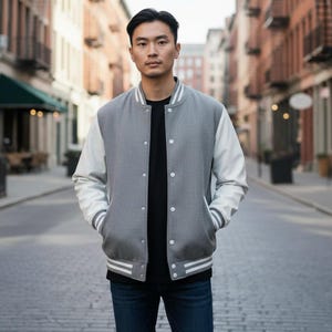 May include: A gray varsity jacket with white sleeves, white stripes on the cuffs and hem, and white buttons. The jacket is worn over a black shirt and blue jeans. The person is standing on a cobblestone street.