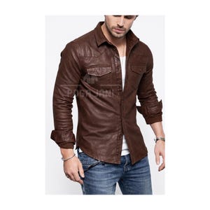 May include: A brown leather shirt with two chest pockets and button fastenings. The long-sleeved shirt has a collared neckline and is worn with blue jeans. The shirt has a rugged, worn appearance.