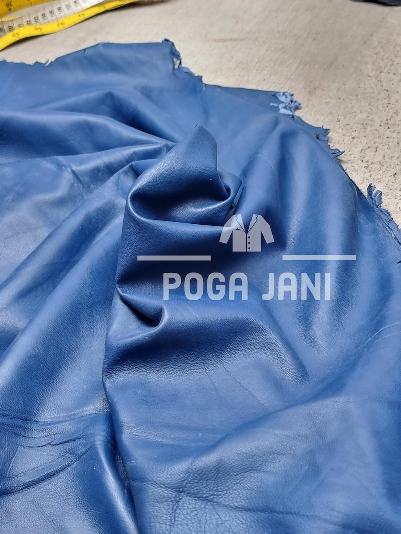 May include: A piece of deep blue leather with visible folds and a textured surface. The text "POGA JANI" is visible in white. A yellow measuring tape is in the background.