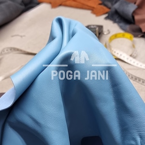May include: A close-up of a piece of light blue leather with the text "POGA JANI". The leather is folded, revealing a lighter underside. A yellow measuring tape and other leather pieces are visible in the background.
