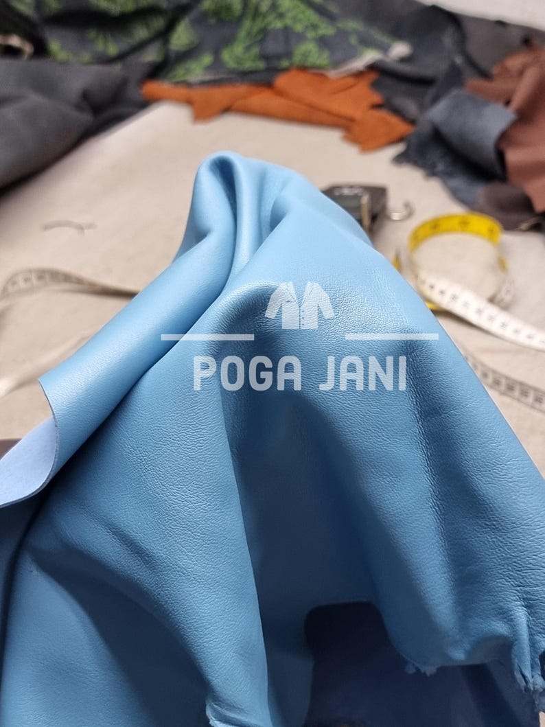 May include: A close-up of a piece of light blue leather with the text "POGA JANI". The leather is folded, revealing a lighter underside. A yellow measuring tape and other leather pieces are visible in the background.