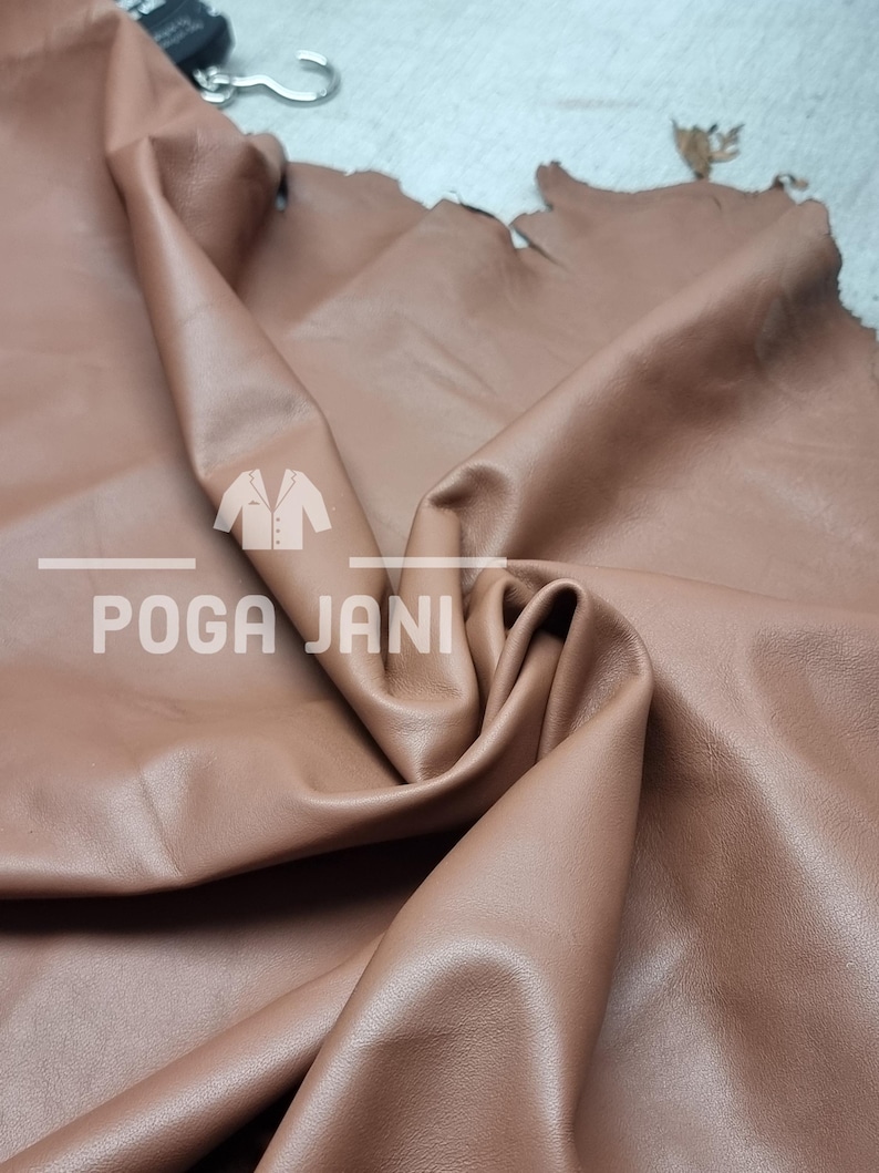 May include: A close-up of a large piece of brown leather with a soft, supple texture. The leather is folded, revealing its thickness and natural grain. The text "POGA JANI" is printed on the leather.