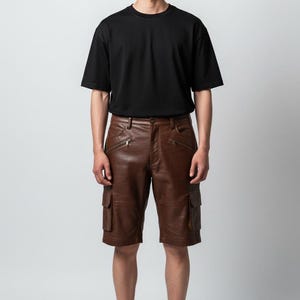 May include: A person wearing a black crew neck t-shirt and brown leather cargo shorts. The shorts have multiple pockets and zipper details. Black sneakers complete the outfit. The background is a plain white.