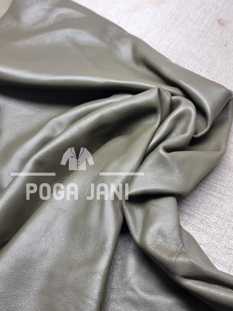 May include: Olive green leather fabric with visible folds and a soft sheen. The fabric is draped, showcasing its texture and flexibility. The text "POGA JANI" is printed on the fabric.