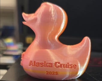 Puede incluir: Figura de pato de goma impresa en 3D en tonos rosa y naranja. El texto "Alaska Cruise 2025" está impreso en el cuerpo del pato. Un artículo de recuerdo.
