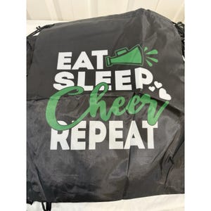 May include: Black drawstring bag with the words "EAT SLEEP CHEER REPEAT" in white and green lettering. A green megaphone graphic is above the word "EAT" and white hearts are to the right of the word "CHEER".
