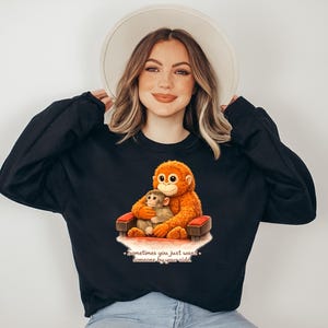 May include: Black sweatshirt featuring a graphic of two monkeys, one orange and one tan, sitting in a red chair. The text "Sometimes you just want someone by your side" is below the graphic.
