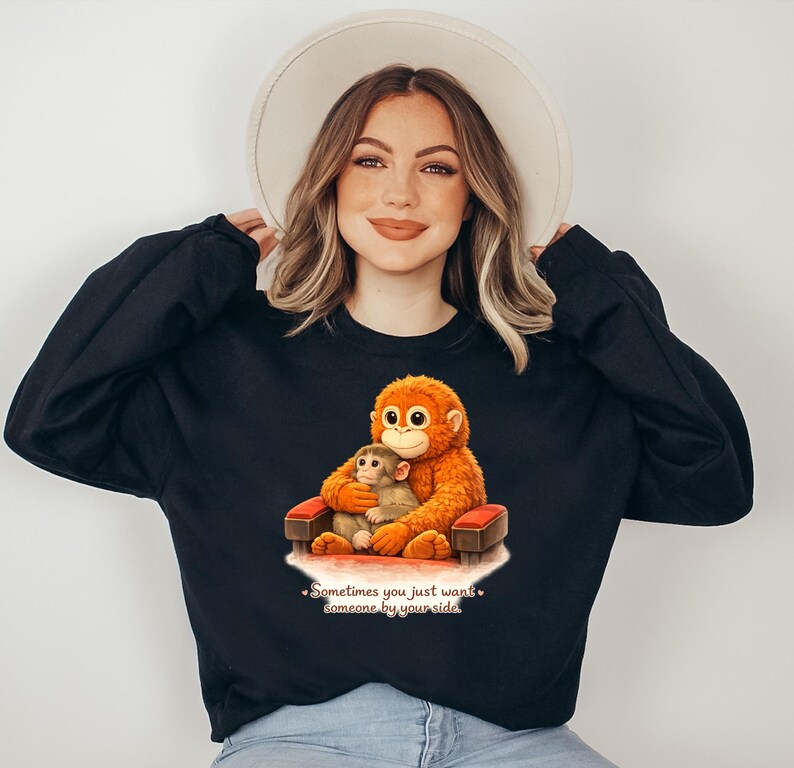 May include: Black sweatshirt featuring a graphic of two monkeys, one orange and one tan, sitting in a red chair. The text "Sometimes you just want someone by your side" is below the graphic.