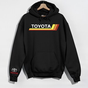 May include: Black hoodie with the word "TOYOTA" in white block letters, above three horizontal stripes in yellow, orange, and red. The Toyota logo is on the left sleeve. The hoodie has a drawstring hood and a front pocket.