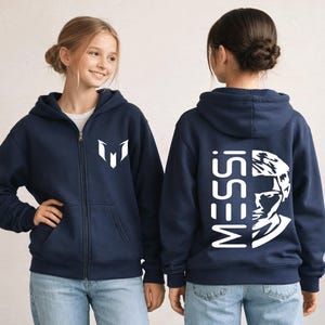 Puede incluir: Una sudadera con capucha y cremallera azul marino que presenta un logotipo blanco en el pecho izquierdo y un gran gr&aacute;fico blanco en la espalda con el texto MESSI junto a un retrato estilizado de un rostro. Incluye capucha y bolsillos.