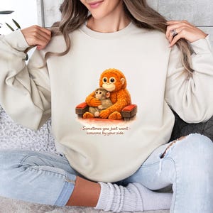 May include: Cream-colored sweatshirt featuring a cartoon orangutan and baby orangutan seated in a small chair. The orangutans are orange and brown. The text "Sometimes you just want someone by your side" is below the image.
