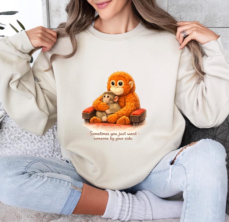 May include: Cream-colored sweatshirt featuring a cartoon orangutan and baby orangutan seated in a small chair. The orangutans are orange and brown. The text "Sometimes you just want someone by your side" is below the image.