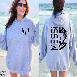 Puede incluir: Una sudadera con capucha gris claro que presenta un logotipo negro en el pecho y un gr&aacute;fico negro grande en la espalda con el texto MESSI junto a un retrato estilizado de una persona. La prenda incluye un bolsillo tipo canguro frontal.
