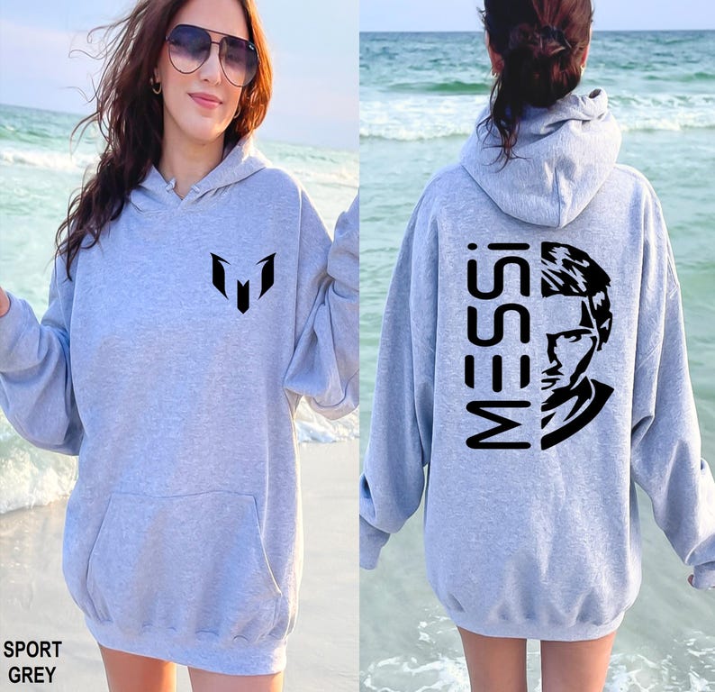 Puede incluir: Una sudadera con capucha gris claro que presenta un logotipo negro en el pecho y un gr&aacute;fico negro grande en la espalda con el texto MESSI junto a un retrato estilizado de una persona. La prenda incluye un bolsillo tipo canguro frontal.