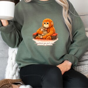 May include: Green sweatshirt featuring a graphic of two orangutans, one embracing the other on a small red bench. The text "Sometimes you just want someone by your side" is below the image. The sweatshirt is a solid green color.