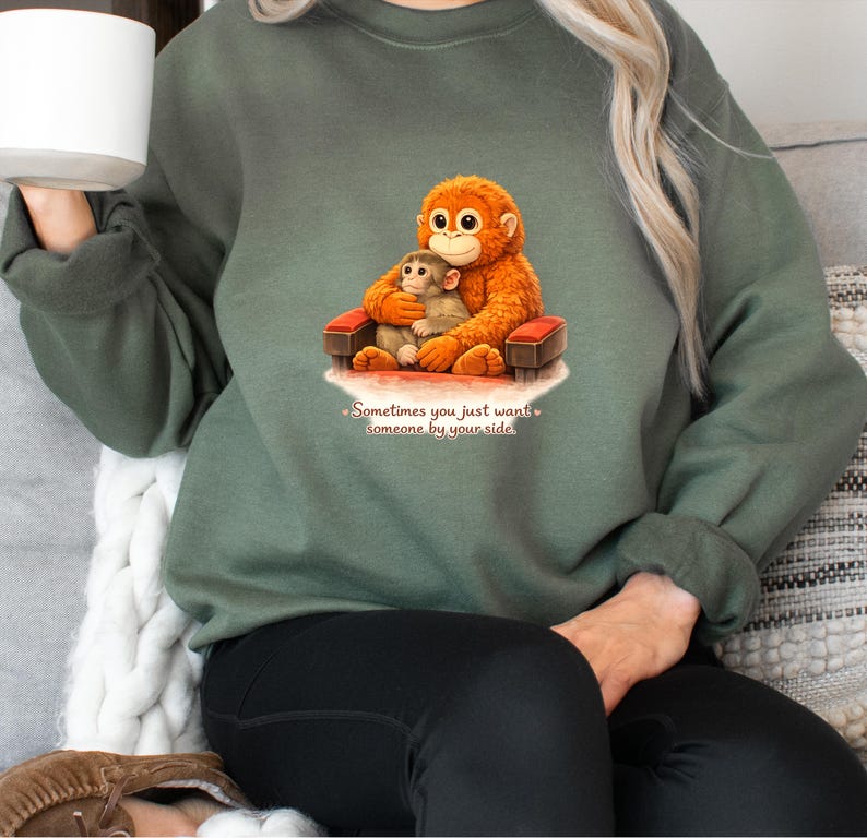 May include: Green sweatshirt featuring a graphic of two orangutans, one embracing the other on a small red bench. The text "Sometimes you just want someone by your side" is below the image. The sweatshirt is a solid green color.