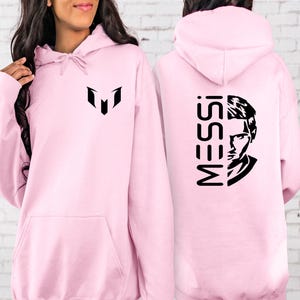 Puede incluir: Una sudadera con capucha de color rosa claro que presenta un logotipo gr&aacute;fico negro en el pecho. La parte posterior de la sudadera muestra el texto MESSI en una fuente negra vertical junto a una ilustraci&oacute;n de retrato en blanco y negro.