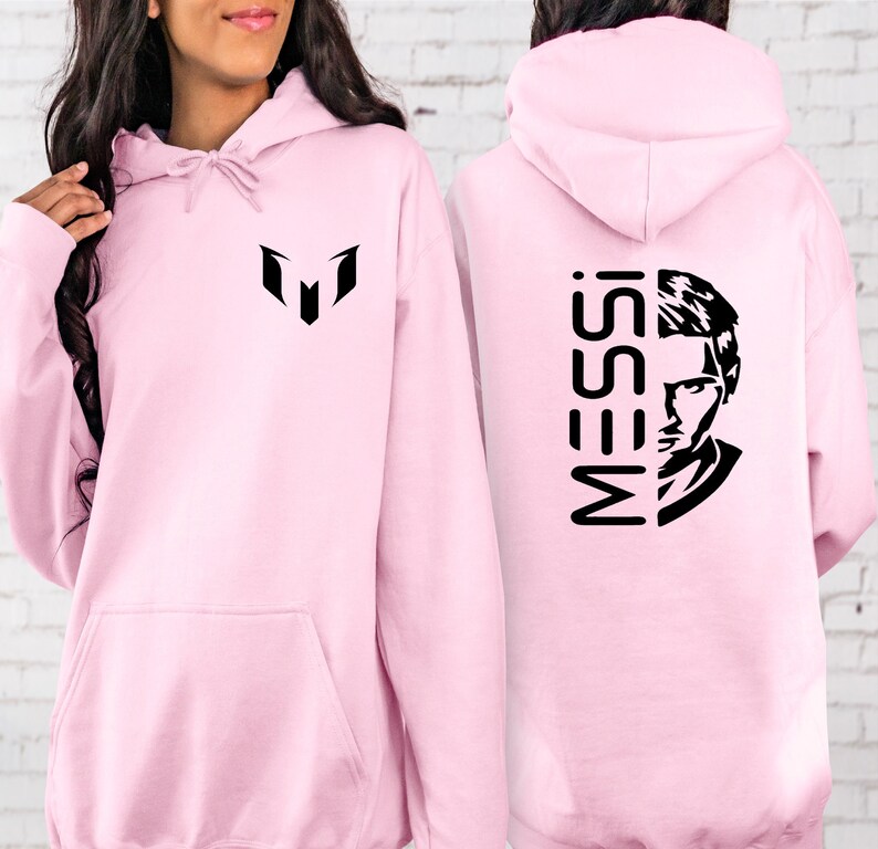 Puede incluir: Una sudadera con capucha de color rosa claro que presenta un logotipo gr&aacute;fico negro en el pecho. La parte posterior de la sudadera muestra el texto MESSI en una fuente negra vertical junto a una ilustraci&oacute;n de retrato en blanco y negro.