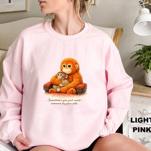 May include: Light pink sweatshirt featuring a cartoon illustration of two orangutans. One large orangutan is hugging a smaller one while seated on a small red and brown chair. The text "Sometimes you just want someone by your side" is below the illustration.