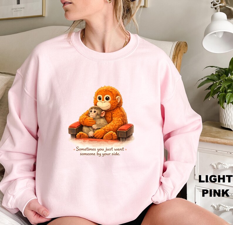 May include: Light pink sweatshirt featuring a cartoon illustration of two orangutans. One large orangutan is hugging a smaller one while seated on a small red and brown chair. The text "Sometimes you just want someone by your side" is below the illustration.