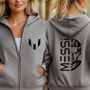 Puede incluir: Una sudadera con capucha y cremallera en color gris jaspeado con un logotipo negro en el pecho izquierdo. La parte posterior muestra el texto MESSI en letras negras grandes junto a una ilustraci&oacute;n gr&aacute;fica negra estilizada de un rostro.