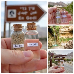 May include: Two small glass bottles with cork stoppers, labeled "Ein Gedi." One contains a beige substance, the other a pink liquid. The bottles are held in a hand, with a sign in the background that reads "En Gedi."