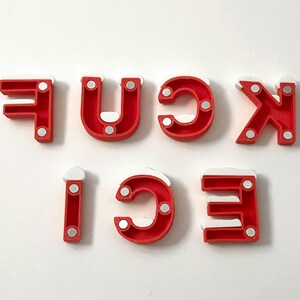 Pu&ograve; includere: Lettere rosse che compongono la parola "FUCK ICE" con accenti bianchi e piccoli magneti argentati. Le lettere sono disposte su uno sfondo bianco. Le lettere sono stampate in 3D.
