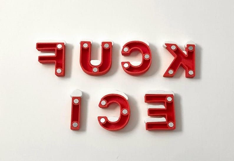 Pu&ograve; includere: Lettere rosse che compongono la parola "FUCK ICE" con accenti bianchi e piccoli magneti argentati. Le lettere sono disposte su uno sfondo bianco. Le lettere sono stampate in 3D.