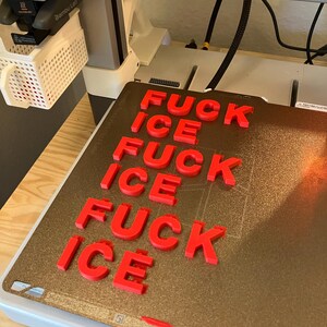 Pu&ograve; includere: Lettere rosse stampate in 3D che compongono "FUCK ICE" su un piano di stampa dorato. Le lettere sono disposte in un motivo ripetuto. La stampante 3D &egrave; visibile sullo sfondo.