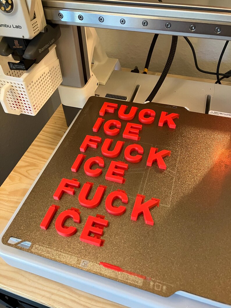 Pu&ograve; includere: Lettere rosse stampate in 3D che compongono "FUCK ICE" su un piano di stampa dorato. Le lettere sono disposte in un motivo ripetuto. La stampante 3D &egrave; visibile sullo sfondo.