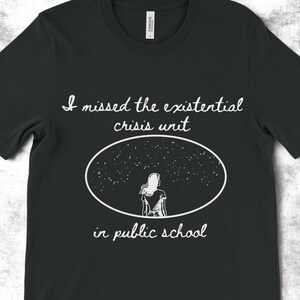 Könnte beinhalten: Schwarzes T-Shirt mit weißem Text, der "Ich habe die existenzielle Kriseneinheit in der öffentlichen Schule vermisst" lautet. Der Text ist in handschriftlicher Form geschrieben. Im Zentrum des Shirts befindet sich eine Silhouette einer Person, die in den sternenklaren Nachthimmel blickt.