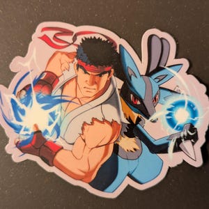 May include: A sticker featuring two characters, one from Street Fighter and the other from Pokemon. The Street Fighter character is a muscular man with a red headband, and the Pokemon character is a blue and black Lucario. Both characters are emitting blue energy.