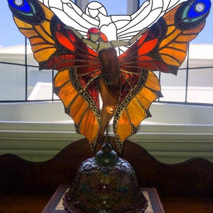 May include: A stained glass butterfly lamp with orange, red, and yellow wings. The lamp features a figure with outstretched arms and is set on a decorative base atop a vintage book. The wings have blue accents.