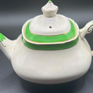 May include: Cream-coloured ceramic teapot with green and gold detailing. The lid and handle have green accents, and the spout is also decorated with green. The teapot has a rounded body and a small lid finial.