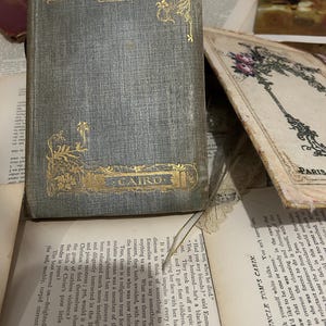 Może przedstawiać: Zabytkowa książka zatytułowana "Medieval Towns" ze złotymi literami i detalami. Książka ma szaro-zieloną okładkę, widoczne jest słowo "Cairo". Inne książki i strony są w tle.