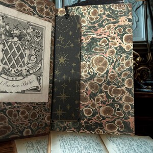 May include: An open antique book with a decorative marbled cover and a black bookmark featuring a gold star pattern. The book's pages are aged, and the bookmark is placed between the pages. The book is on a wooden surface.