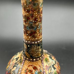 May include: A decorative, ornate vase with a narrow neck and bulbous base. The vase features a detailed floral design in shades of blue, red, and gold on a tan background. The neck has a textured, dotted pattern.