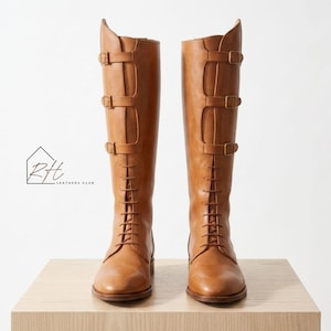Könnte beinhalten: Ein Paar hohe, hellbraune Lederreitstiefel. Die Stiefel haben eine Schnürung vorne, drei Schnallenriemen und einen hohen Schaft. Die Stiefel werden auf einer hellen Holzoberfläche vor weißem Hintergrund präsentiert. Das Logo "Rtt Leathers Club" ist sichtbar.