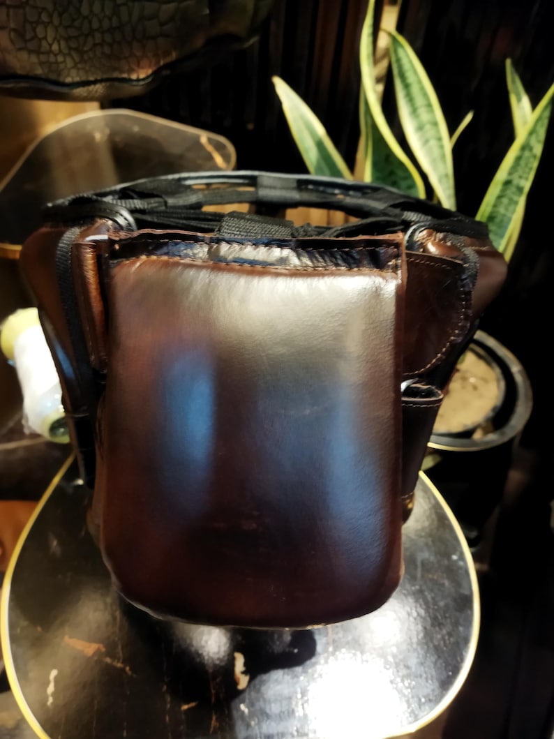May include: A brown leather boxing headguard with black straps and a padded interior. The headguard is designed to protect the head and face during boxing training.