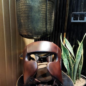 May include: A brown leather boxing headguard with padding around the ears and forehead. The headguard is sitting on a black and gold table.