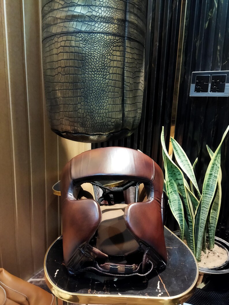 May include: A brown leather boxing headguard with padding around the ears and forehead. The headguard is sitting on a black and gold table.
