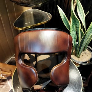 May include: A brown leather boxing headgear with padding around the ears and forehead. The headgear is designed to protect the head during boxing training.