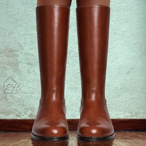May include: A pair of tall, brown leather riding boots. The boots are knee-high with a smooth, polished finish. The image shows the boots from the ankles up, with a light green background and a logo in the corner.